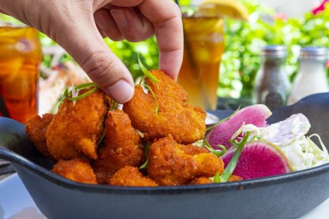 Hand placing crispy fried chicken pieces in a black skillet with fresh vegetables and condiments.