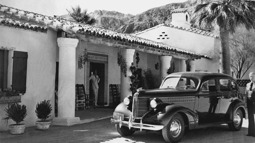 Vintage black car parked outside a Spanish-style resort building.