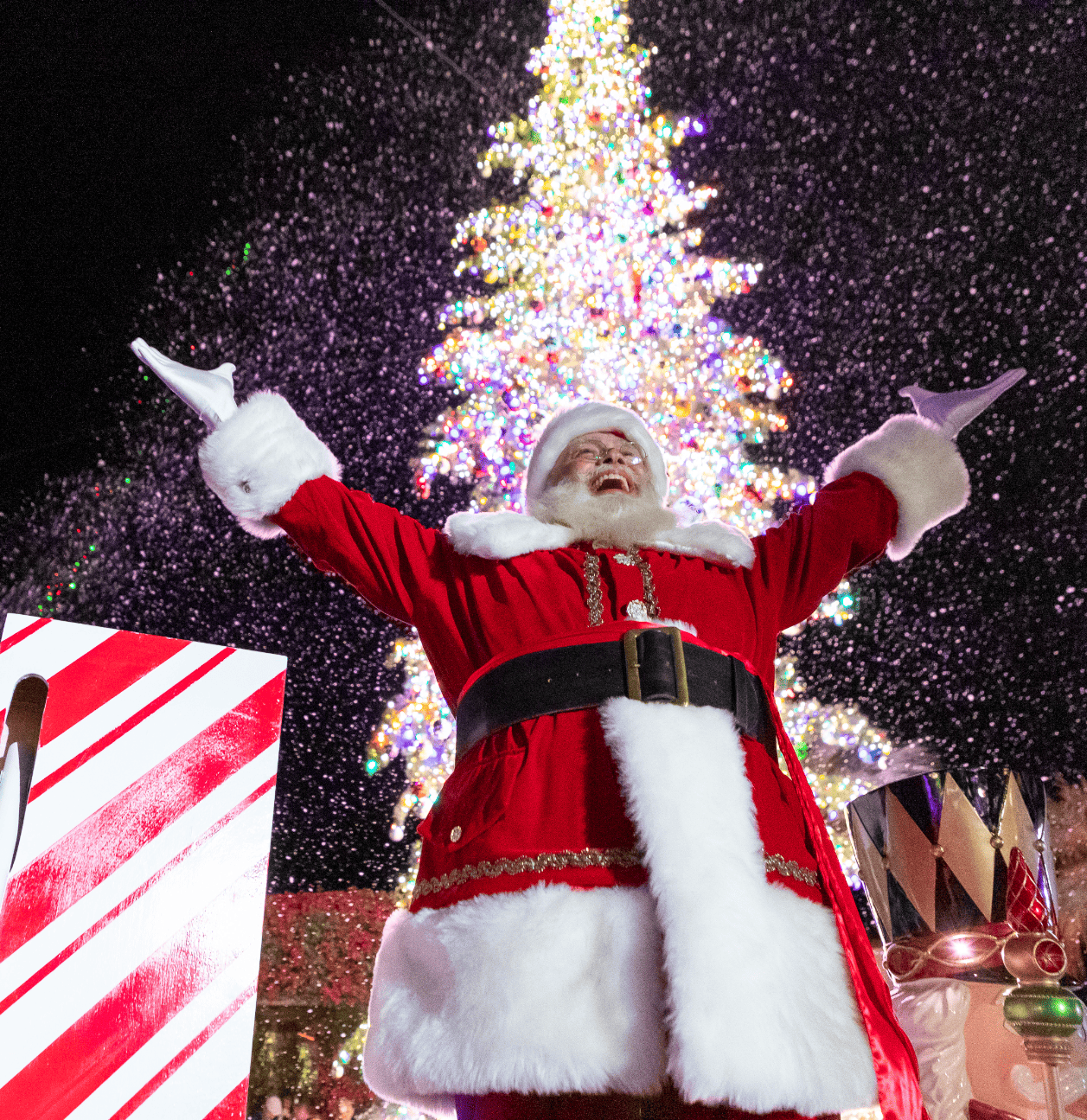 Santa Claus with arms raised in front of a decorated Christmas tree.