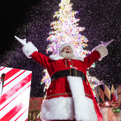 Santa Claus with arms raised in front of a decorated Christmas tree.