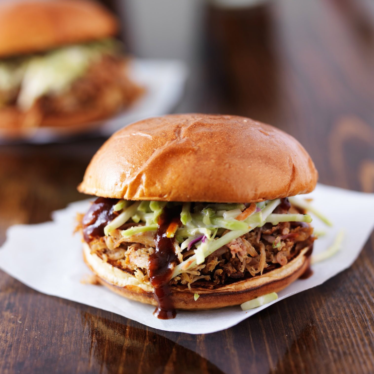 Pulled pork sandwich with coleslaw and barbecue sauce on a bun, served on a wooden table.